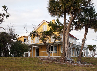 [Atop a knoll is a large three-storied yellow-sided Victorian-inspired house with white trim and open white wooden porches on the visible sides of the building and on both the first and second level (open and not covered porches on the second level). Several cedar and palm trees in the foreground.]